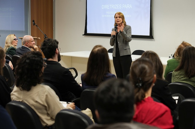 Mulher palestrante no palco com microfone, diante de público sentado, e tela de projeção com título "DIRETRIZES PARA" em ambiente de seminário.