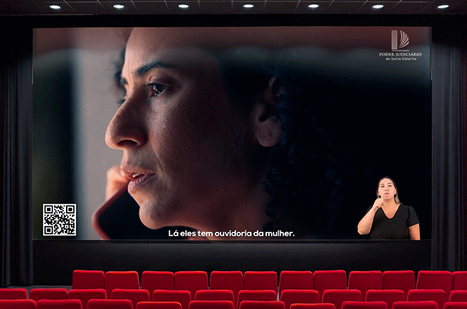A imagem mostra uma tela de cinema com uma mulher preocupada, em chamada telefônica.Um texto sobre ouvidoria da mulher, QR code e intérprete de libras à frente.