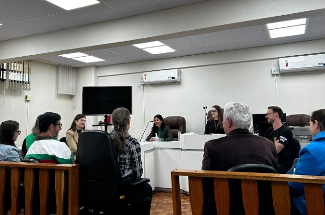 A imagem mostra uma sala de audiências onde há uma mulher sentada na mesa e outras pessoas sentadas em bancos à sua frente. A sala é clara e simples, há um televisor, paredes brancas e piso claro.