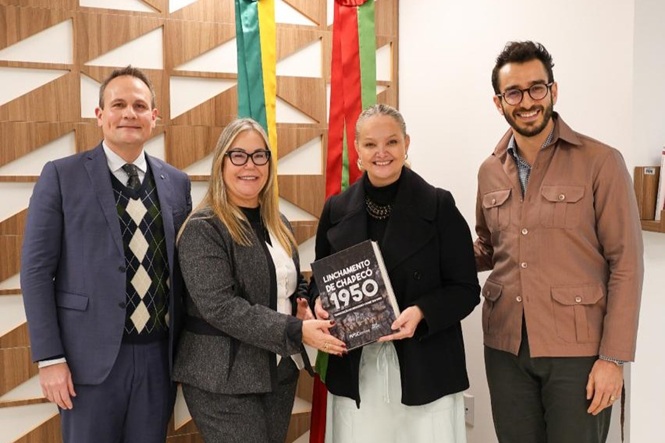 Quatro pessoas posam formalmente, duas segurando o livro "Lançamento de Chapecó 1950", com bandeiras e painel de madeira ao fundo.