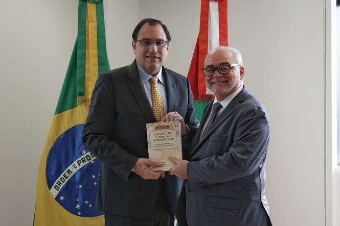 Foto horizontal: Francisco Oliveira Neto recebe livro de autoridade diante das bandeiras do Brasil e SC.