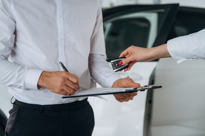 Na imagem, vemos duas pessoas parcialmente. A pessoa à esquerda está segurando uma prancheta e assinando um documento. A pessoa à direita está passando uma chave de carro para a outra.