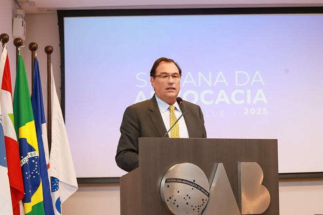 O Presidente do TJSC, está em um pulpito com as iniciais: OAB, durante uma cerimônia. Várias bandeiras estão em postes ao fundo. Há um projetor com o texto "Semana da Advocacia 2025" na tela.