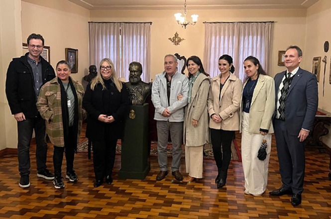 A imagem mostra oito pessoas posando sorridentes ao redor de um busto em uma sala decorada com piso de madeira, lustre e pinturas, transmitindo um clima descontraído em ambiente de museu ou casa histórica.