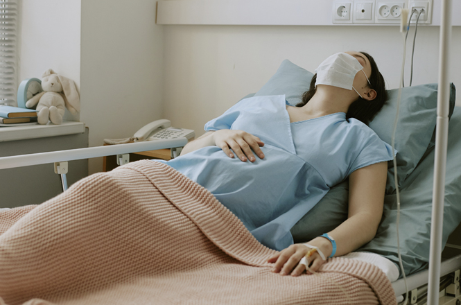 A imagem mostra uma mulher deitada em uma cama de hospital, usando camisola azul e máscara facial, com pulseira de identificação no pulso e mão apoiada sobre o abdômen.