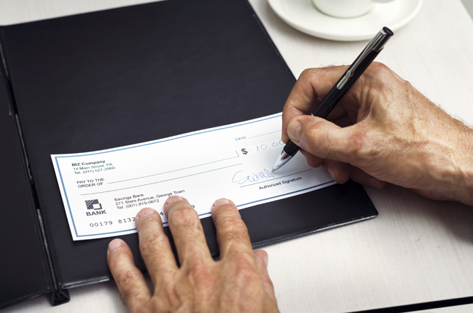 A imagem mostra uma pessoa assinando um cheque de R$10,00, com a mão direita segurando a caneta e a esquerda apoiando o papel dentro de uma capa preta.