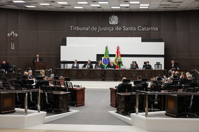 A imagem retrata um ambiente formal e oficial, possivelmente uma sala de tribunal dentro do "Tribunal de Justiça de Santa Catarina", como indicado no painel de fundo.