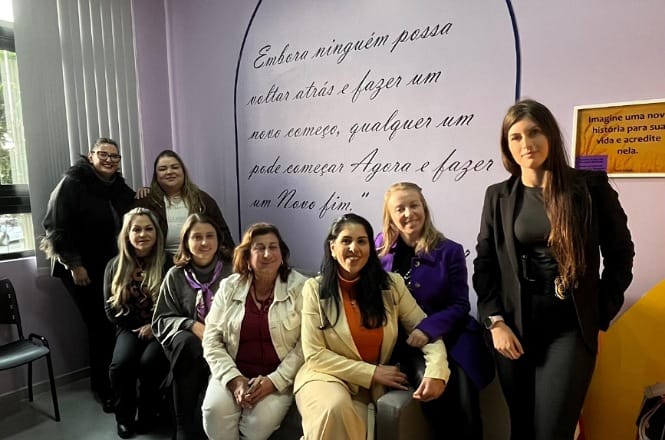 A imagem mostra oito mulheres posando juntas em um ambiente interno, diante de uma parede lilás com frases motivacionais, transmitindo união, positividade e empoderamento coletivo.