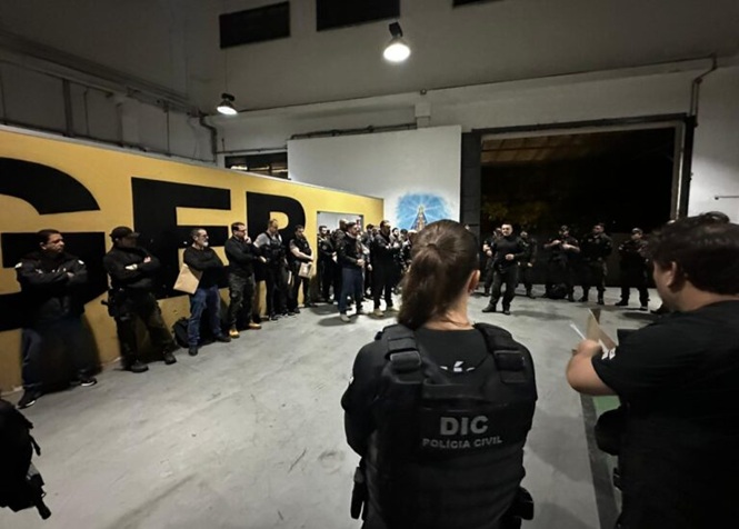 A imagem mostra policiais reunidos em um espaço fechado, com um agente da DIC de costas em primeiro plano, em um briefing diante de uma parede amarela com letras grandes, em clima sério e concentrado.
