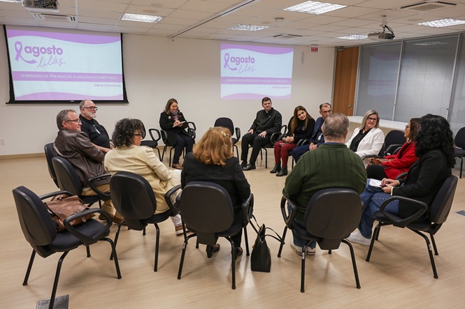 Sala de conferências com pessoas em círculo, engajadas em discussão sobre prevenção à violência doméstica. Ambiente formal e iluminado, marcado por diálogo, cooperação e aprendizado.