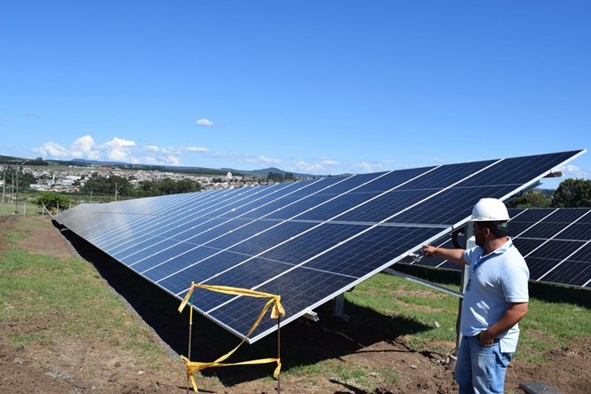A imagem mostra um cenário ao ar livre com um conjunto de painéis solares no primeiro plano. No fundo, é possível ver uma paisagem urbana e algumas colinas ao longe.