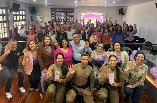 Um grupo de pessoas, incluindo policiais uniformizados, posam para foto em uma sala de conferências, segurando folhetos e acenando para a câmera.