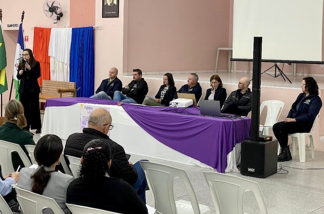 A imagem retrata um evento em um salão, com público sentado assistindo. No palco, uma mulher fala ao microfone diante de uma mesa com toalhas branca e roxa e de um painel com cortinas azul, branca e vermelha.
