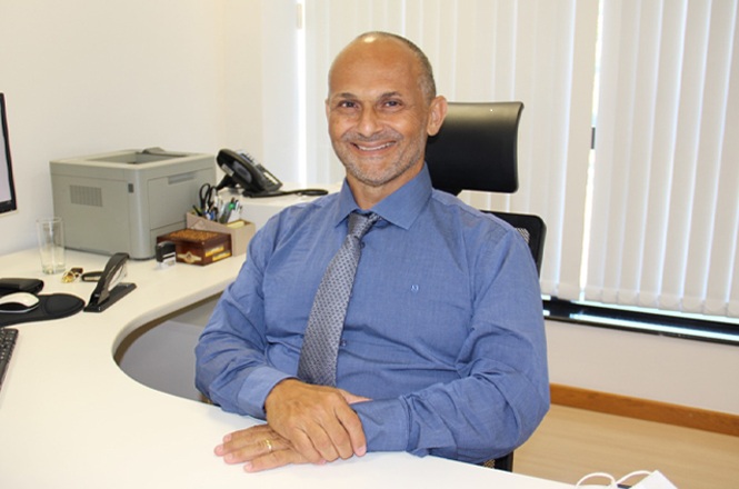 A imagem mostra o magistrado Maurício Cavallazzi Póvoas sentado em uma mesa de escritório, sorrindo, cercado por computador, telefone e materiais de escritório.