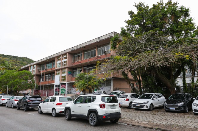 A imagem mostra o interior frontal de um carro estacionado, com volante à esquerda, painel multimídia e detalhes esportivos, enquanto pelo para-brisa é possível ver um estacionamento urbano ao fundo.