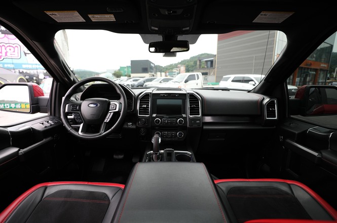A imagem mostra o interior frontal de um carro estacionado, com volante à esquerda, painel multimídia e detalhes esportivos, enquanto pelo para-brisa é possível ver um estacionamento urbano ao fundo.