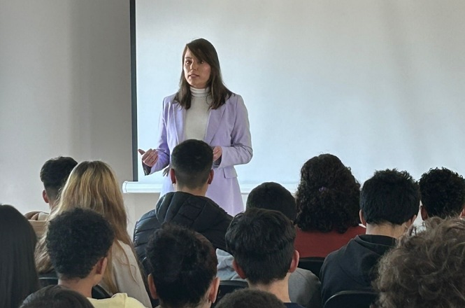 A imagem mostra uma mulher de casaco lilás realizando uma apresentação para uma audiência sentada em um ambiente interno, possivelmente uma sala de aula ou auditório, com atmosfera formal e atenta.