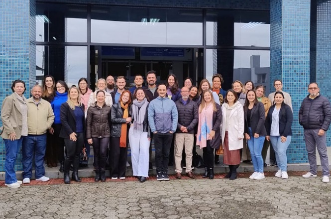 A imagem mostra cerca de trinta pessoas sorrindo e posando em frente a um prédio de azulejos azuis, transmitindo cordialidade e alegria em um ambiente institucional durante o dia.