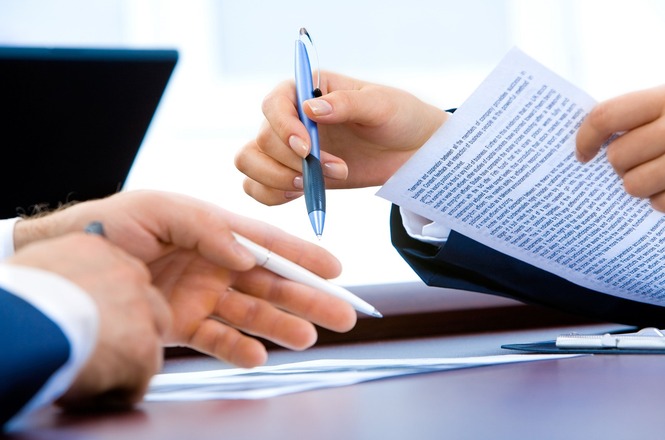 A imagem mostra duas pessoas em um escritório revisando ou assinando um documento sobre uma mesa, com laptop ao fundo, em um ambiente profissional e colaborativo.