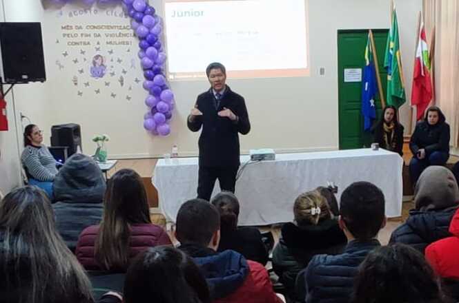 A imagem mostra um palestrante em uma sala de conferências, com público atento, projetor, balões roxos e bandeiras, em um evento formal sobre conscientização contra a violência às mulheres.