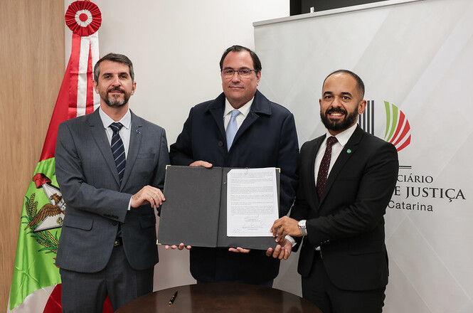 A imagem mostra três homens de terno exibindo um documento em um ambiente institucional, sorrindo para a câmera em um momento formal e profissional