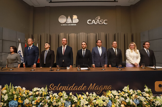 Foto colorida: autoridades alinhadas atrás de mesa solene, auditório institucional, solenidade magna, acompanhando ato em azul, dourado e cinza
