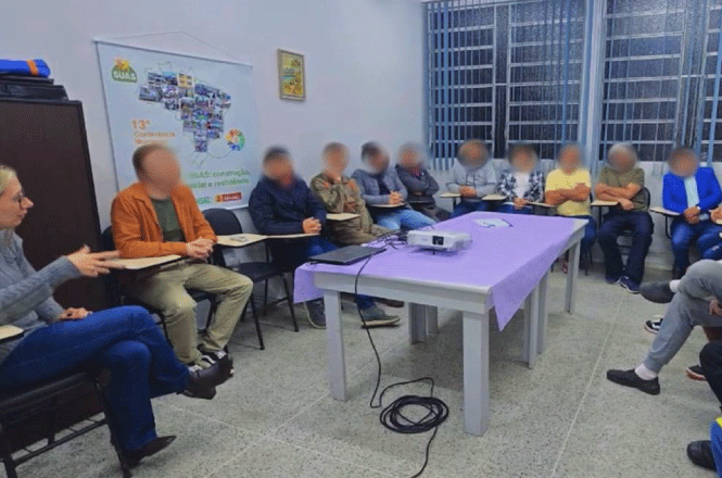 A imagem mostra uma reunião em círculo, com pessoas atentas e engajadas ao redor de uma mesa roxa com projetor e notebook, em uma sala bem iluminada.
