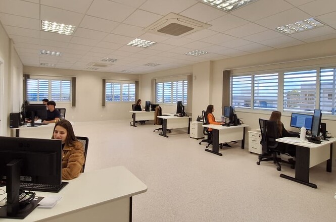 A imagem mostra um escritório moderno e bem iluminado, com pessoas concentradas em seus computadores em mesas brancas, em um ambiente profissional e produtivo.
