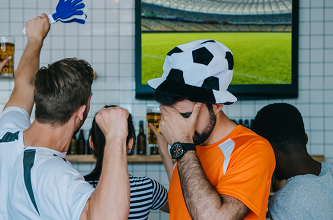 A imagem mostra pessoas em um bar assistindo a um jogo de futebol na TV, com uma delas cobrindo o rosto em frustração, enquanto outras comemoram com uma mão gigante de espuma azul.