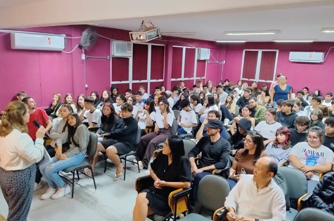 Foto colorida: público reunido em auditório escolar, sala interna ampla, ação educativa, participando de palestra em rosa, branco e cinza