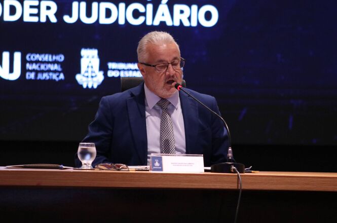 Foto horizontal: desembargador Sidney Eloy Dalabrida fala ao microfone em mesa oficial, painel do Judiciário ao fundo, tons escuros.