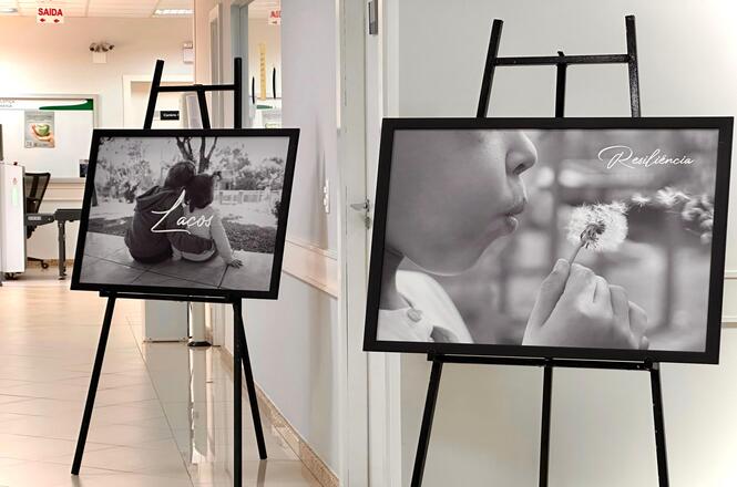 Foto colorida: quadros fotográficos em cavaletes, corredor institucional interno, exposição temática, destacando laços e resiliência em preto e branco
