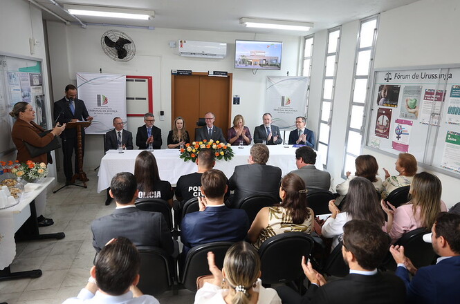 Foto colorida: autoridades à mesa e público aplaudindo, sala do fórum, cerimônia institucional, celebrando ato em branco, bege e verde