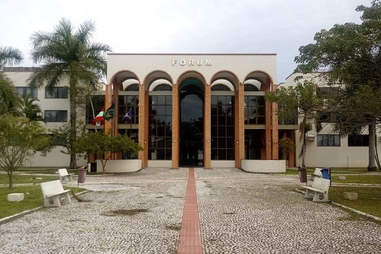Foto colorida: prédio do fórum com arcos na fachada, área urbana ajardinada, sede judiciária, destacando acesso em branco, bege e verde