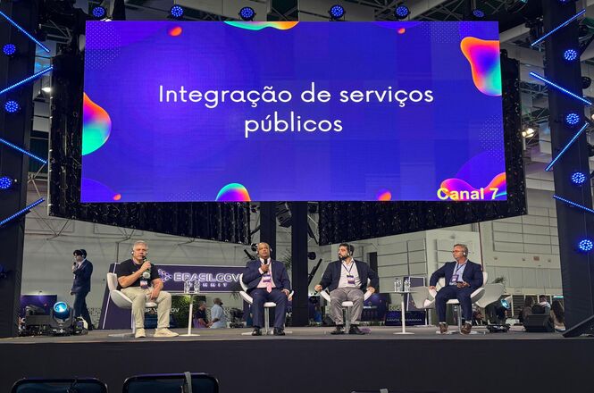 Foto colorida: painel de autoridades em palco, auditório de evento nacional, integração de serviços públicos, debatendo tema em roxo, azul e branco