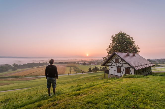 Foto colorida: pessoa observa o pôr do sol em campo verde, casa rural ao lado, paisagem campestre, contemplando horizonte em verde, rosa e dourado