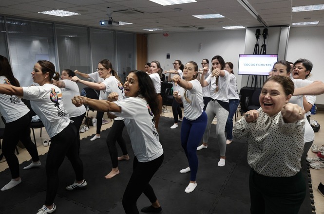 Foto horizontal: A desembargadora Hildemar Meneguzzi, de blusa de bolinhas, sorrindo pra câmera, com servidoras do judiciário treinando defesa pessoal
