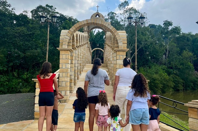 Foto colorida: grupo caminhando sobre ponte de pedra, parque arborizado, lazer ao ar livre, atravessando em tons bege, verde e azul