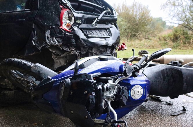 Foto colorida: motocicleta caída e carro danificado na via, área rural externa, acidente de trânsito, evidenciando colisão em azul, preto e cinza