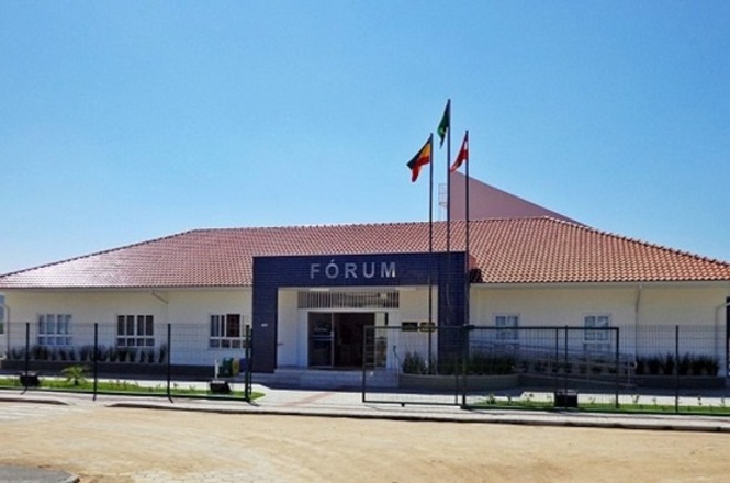 Foto colorida: prédio do fórum com bandeiras hasteadas, área urbana ajardinada, sede judiciária, destacando fachada em branco, azul e verde
