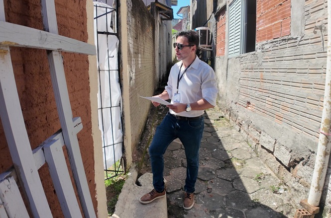 Foto colorida: oficial de justiça em viela urbana, edificações laterais, diligência externa, conferindo documentos em bege, branco e cinza