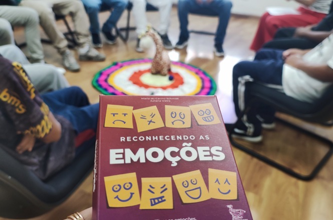 Foto colorida: livro “Reconhecendo as Emoções” em primeiro plano, roda de pessoas sentadas, prática restaurativa, mediando diálogo em tons coloridos