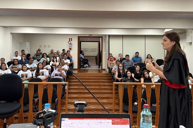 Fotografia horizontal:sala de audiência com piso de madeira e cadeiras escuras, sessão de julgamento, advogada realiza defesa oral gesticulando, em tons marrom, preto e branco