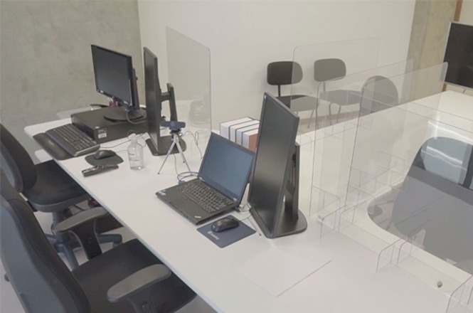 Foto colorida: mesa de atendimento com computadores e divisórias acrílicas, sala interna, audiência de custódia, organizando trabalho em branco, preto e cinza