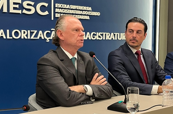 Fotografia horizontal: Rubens Schulz fala ao microfone enquanto outro magistrado observa, sala institucional com fundo azul e texto branco, evento jurídico em tons azul, cinza e branco