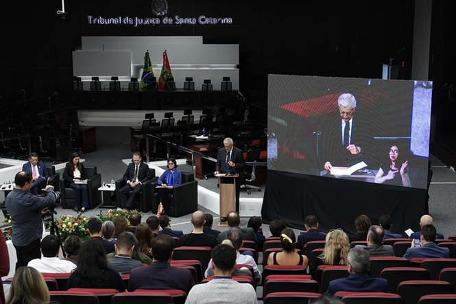 Foto colorida: plenário do TJSC, com público e painel, ambiente institucional, evento jurídico, acompanhando palestra em preto, vermelho e cinza