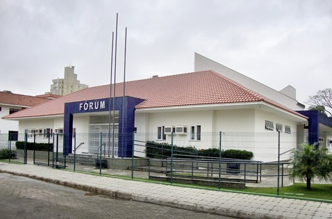 Foto colorida: prédio do fórum com bandeiras hasteadas, área urbana ajardinada, sede judiciária, destacando fachada em branco, azul e verde