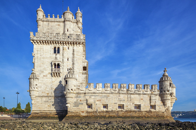 Foto colorida: Castelo medieval europeu de pedra à beira‑mar, paisagem costeira, patrimônio arquitetônico, destacando estrutura em bege, azul e cinza