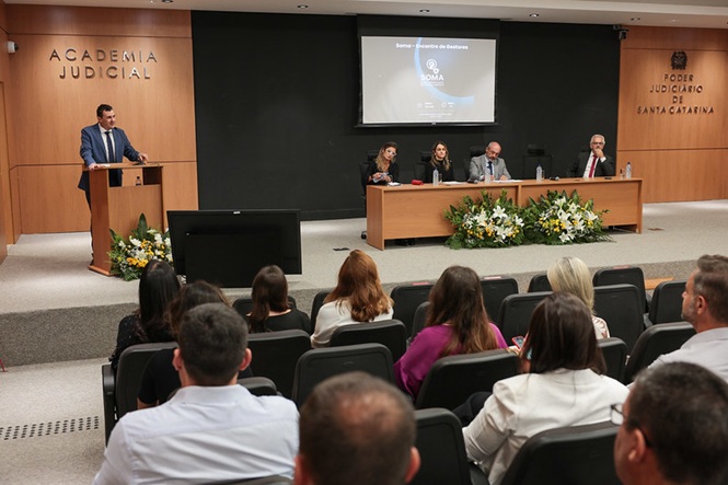 Fotografia horizontal: palestrante, mesa e público em auditório com palco, telão e flores em evento institucional, homem discursa enquanto mesa acompanha e plateia assiste, em tons bege, preto e verde