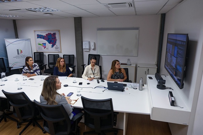 Foto horizontal: mulheres participam de videoconferência em sala do TJSC; tela exibe reunião; ambiente institucional; tons branco, preto e azul.